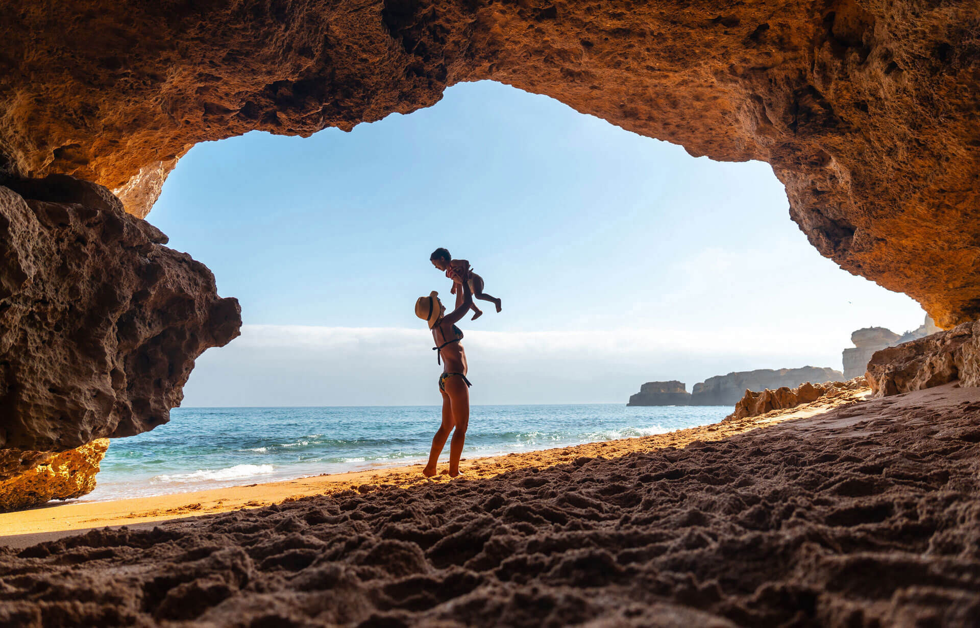 Blickwinkel vom Land durch eine Grotte Richtung Meer. Eine junge Frau hält ihr kleines Kind hoch.