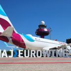 Ein Flugzeug der Eurowings steht auf dem Vorfeld. Vor dem Flugzeug bilden weiße Buchstaben den Schriftzug "HAJ > EUROWINGS". Im Hintergrund ist der Tower in Eurowingsfarben Burgundy bemalt zu sehen.