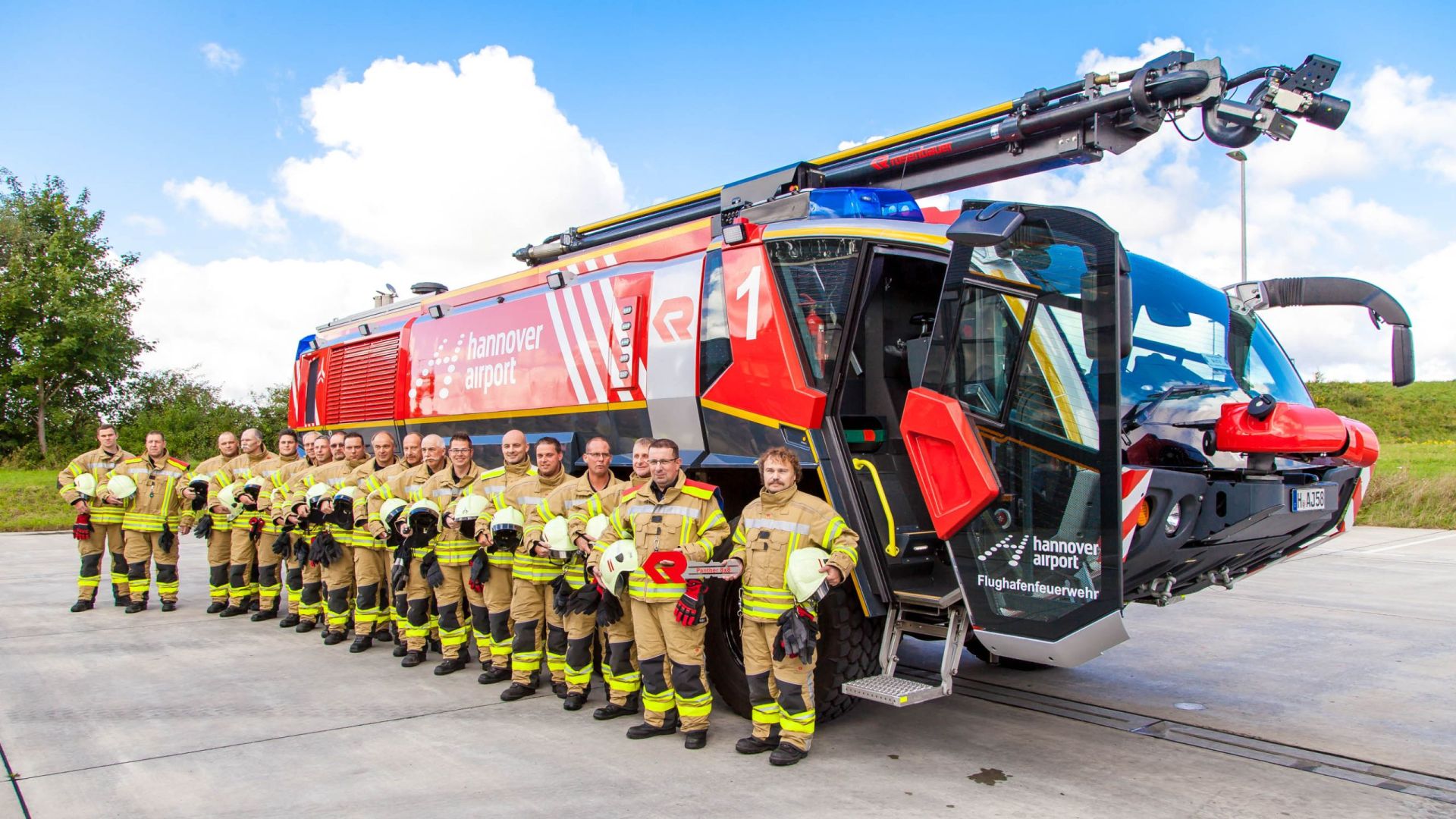 Mitarbeiter der Flughafenfeuerwehr stehen in der neuen Dienstkleidung aufgereiht vor einem Feuerwehrfahrzeug.