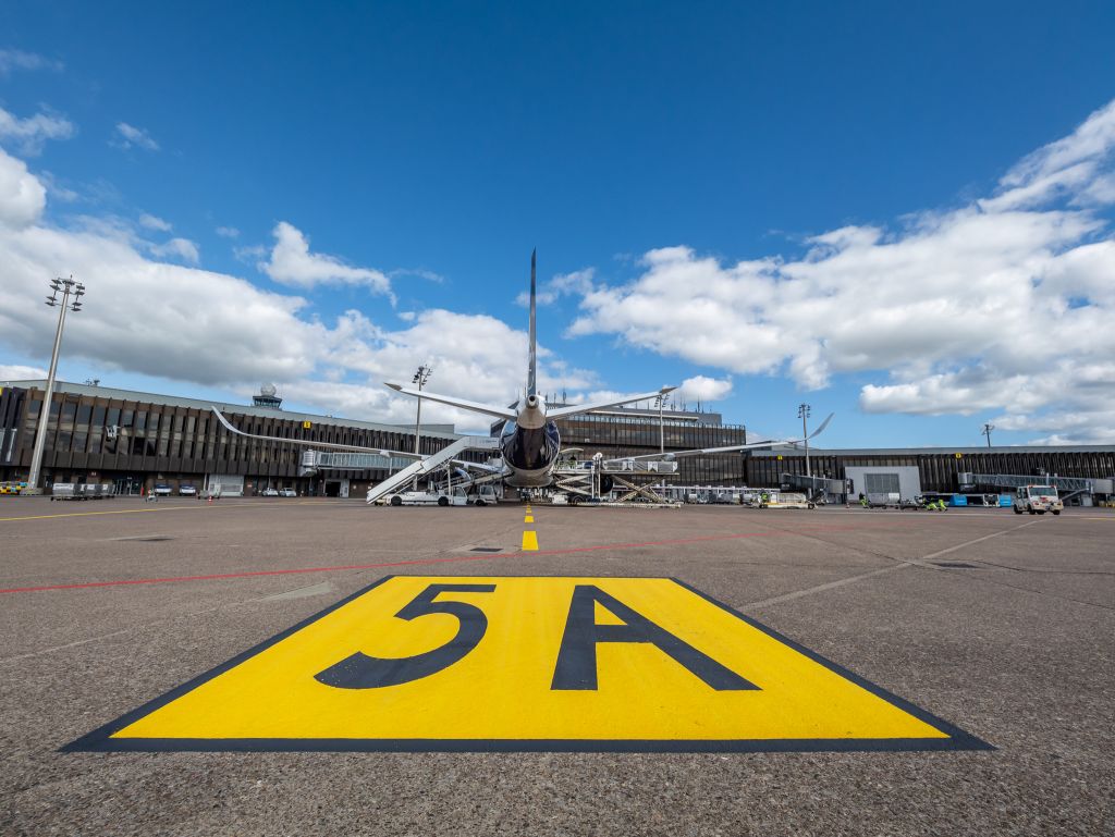 Ein Flugzeug der Lufthansa wird am Hannover Airport ent- und beladen. Man blickt von hinten auf das Flugzeug, dass am Terminal steht. Im Vordergrund des Bildes ist die markant gelbe Positionsmarkierung 5 A.