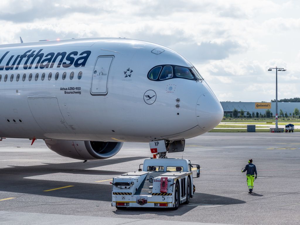 Ein pushback-Fahrzeug drückt das Flugzeug zurück. Ein Mitarbeiter begleitet zu Fuß den Abfertigungsprozess und ist per Headset mit dem Piloten verbunden.