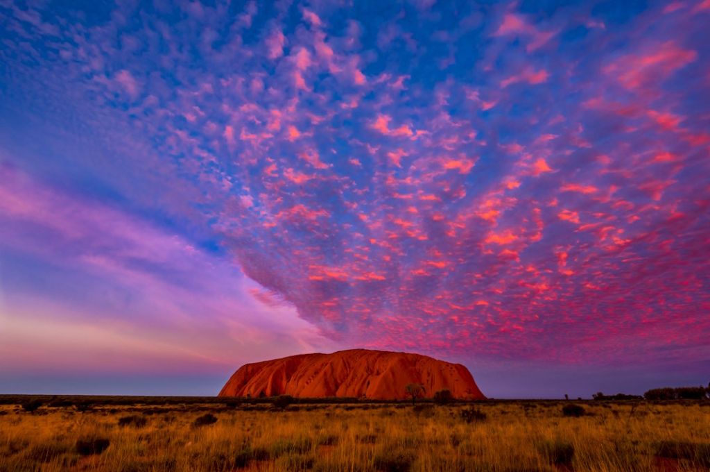 Der Ayers Rock strahlt rot in der Abendsonne. Am Himmel sieht man ebefalls rot angestrahlte Wolken der untergehenden Sonne.