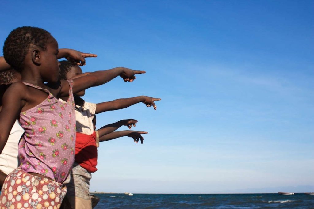 Afrikanische Kinder stehen am Meer und zeigen mit ihren Händen auf das Wasser hinaus.