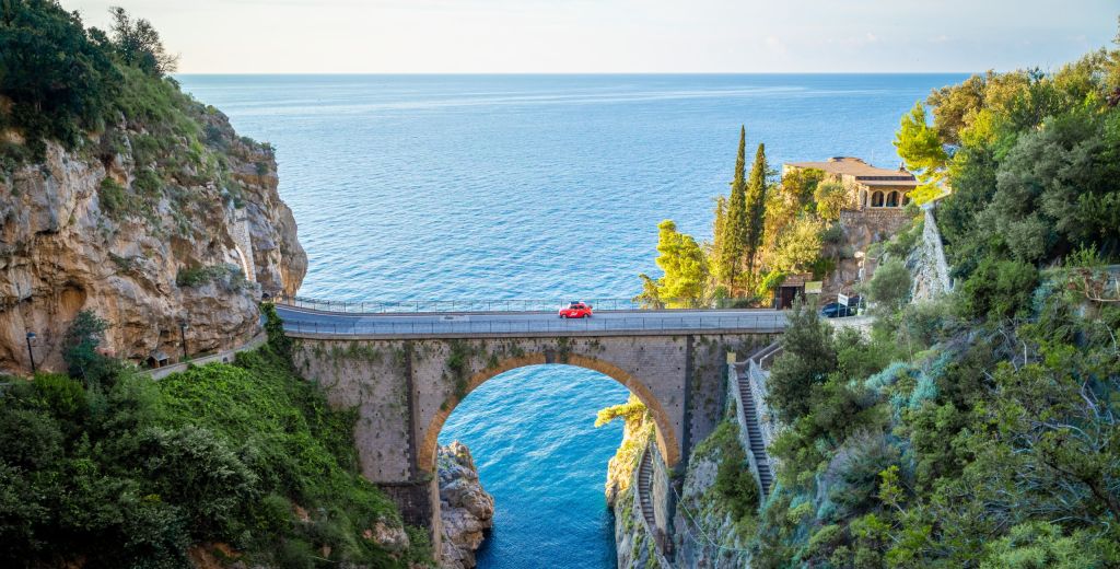 Ein kleiner, roter Fiat 500 überquert eine hohe Brücke. Man sieht unter und hinter der Brücke das blaue Meer.