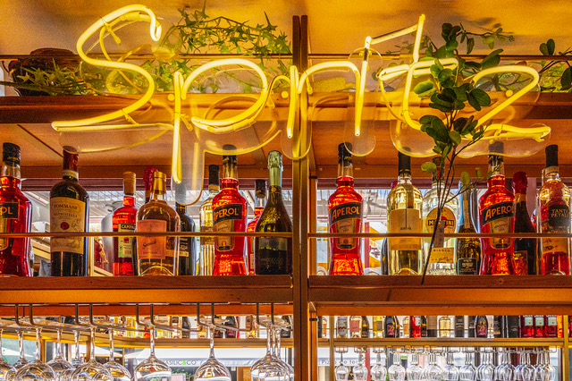 Blick auf die Rückwand einer Bar mit gelbem Neonschriftzug Spritz und ausgeleuchteten Flaschen und Gläsern.