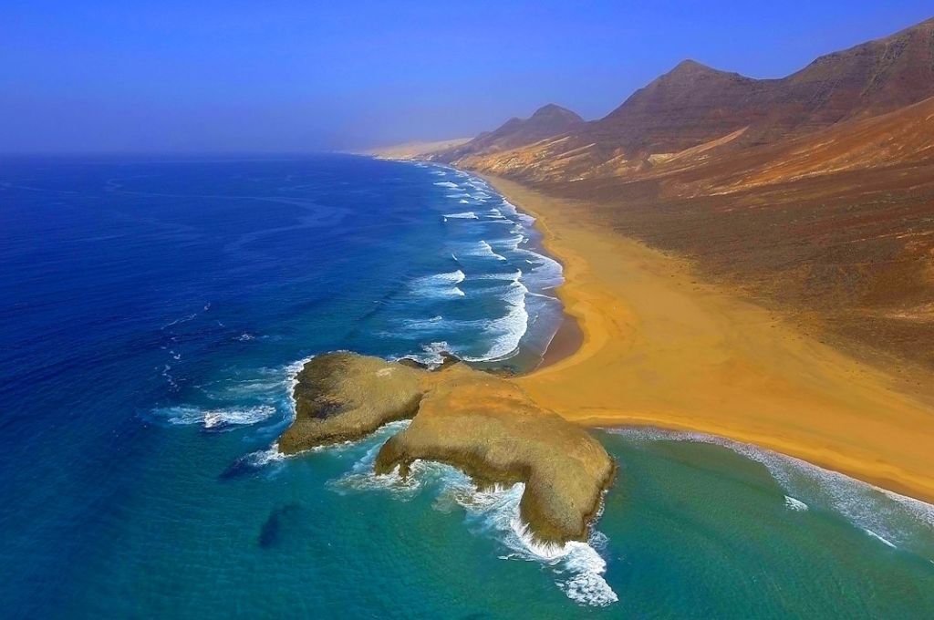 Der Blick von oben die Küste einer der kanarischen Inseln. Meereswellen treffen auf einen langen Sandstrand, an den sich eine Bergkette anschließt.